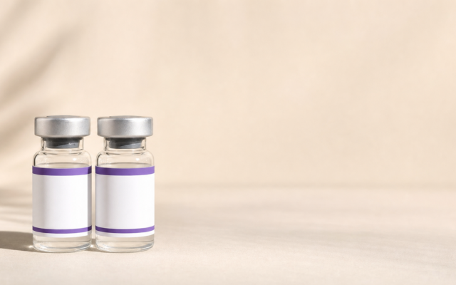 Two small glass medication vials with silver caps and blank white labels featuring purple borders, sitting side by side on a neutral beige surface.